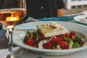 Salad with feta cheese and olives in a white bowl beside a glass of amber wine