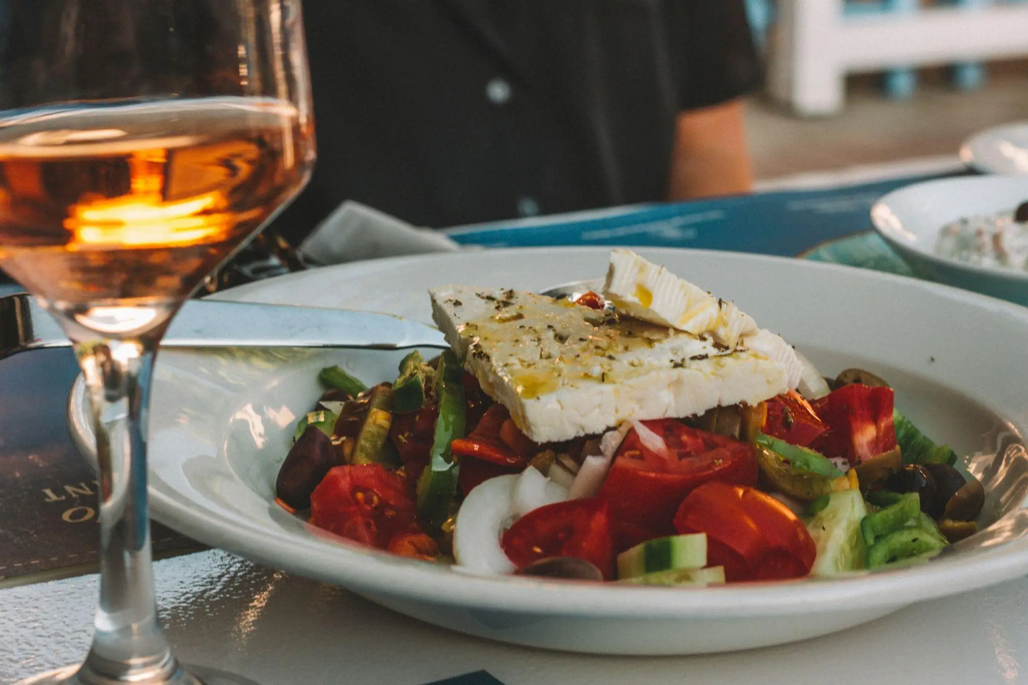 Salad with feta cheese and olives in a white bowl beside a glass of amber wine