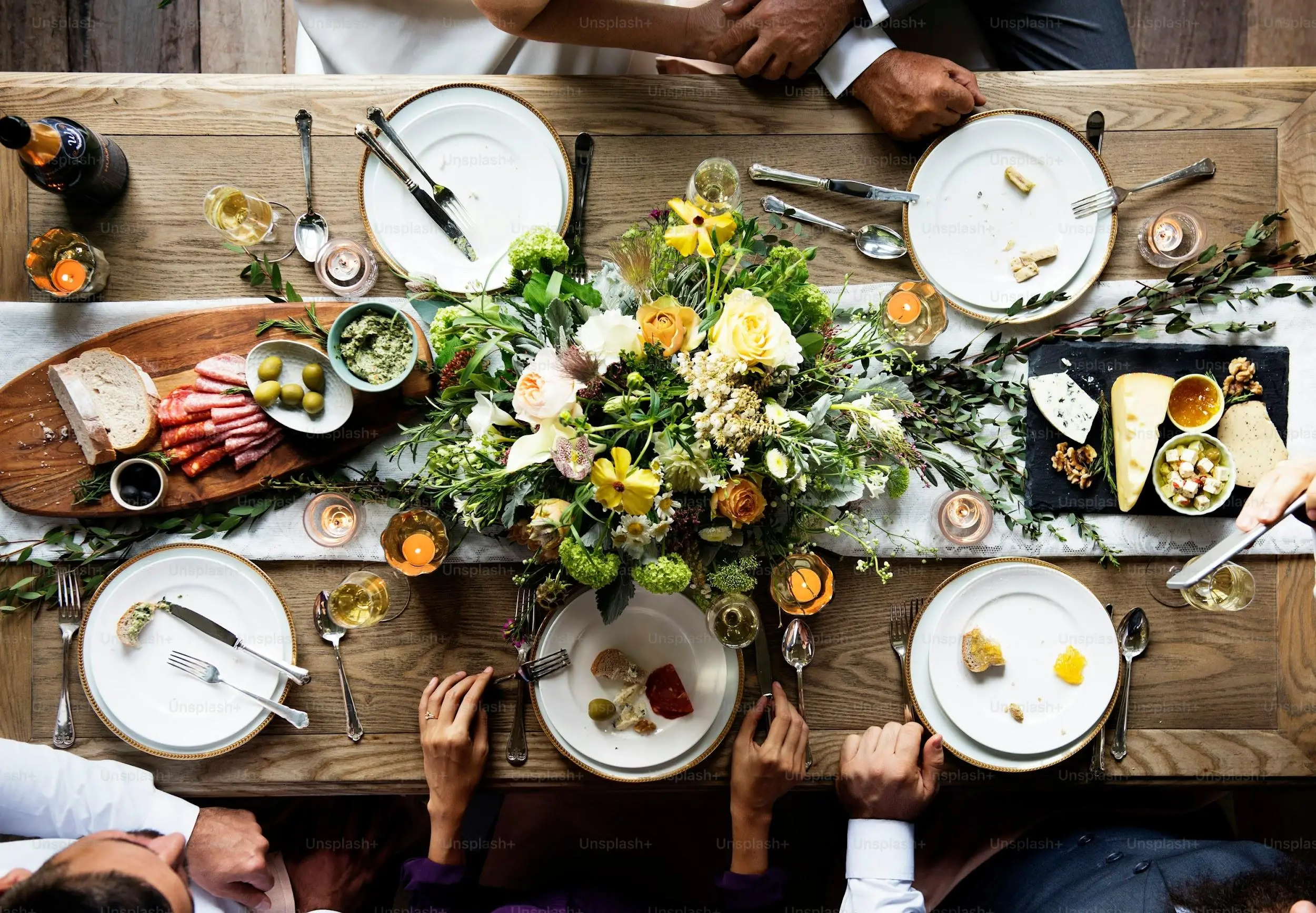 Dining table with charcuterie, cheeses, and floral centerpiece surrounded by people in elegant setting