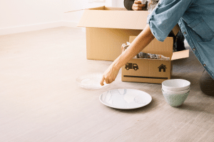 Person unpacking dishes from cardboard box on wooden floor in bright room