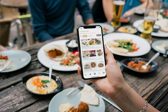 Hand holding smartphone displaying food delivery app at outdoor dining table with various dishes