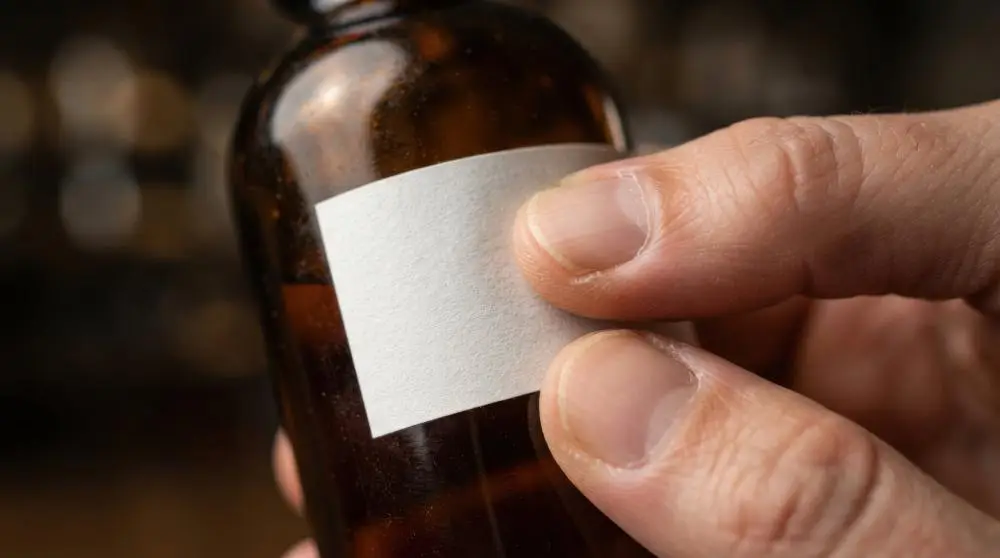 Hand holding small brown glass bottle with blank white label in warm lighting.
