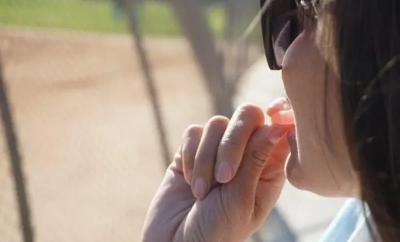 Person wearing sunglasses eating candy near a baseball field on a sunny day