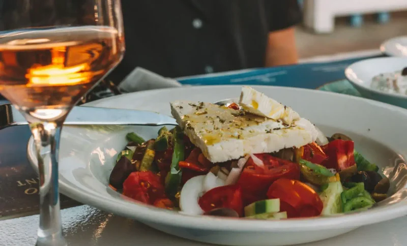 Salad with feta cheese and olives in a white bowl beside a glass of amber wine