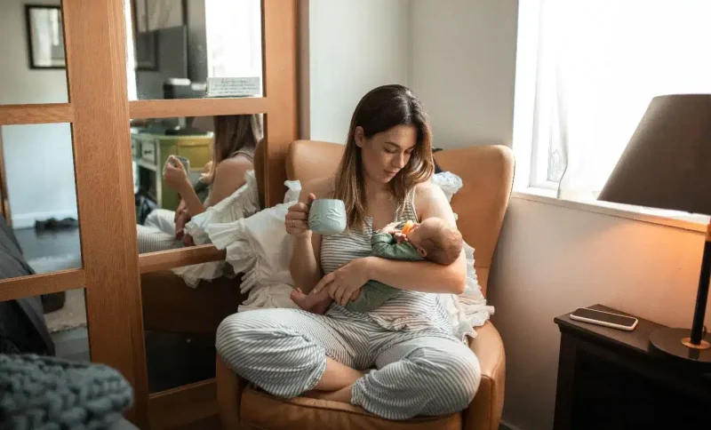 Woman in striped pajamas holding baby and mug in cozy living room chair