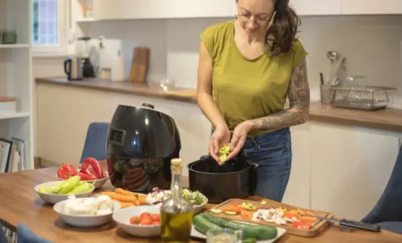 Person preparing vegetables in air fryer on wooden kitchen counter with fresh produce and olive oil
