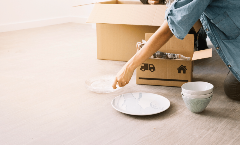Person unpacking dishes from cardboard box on wooden floor in bright room
