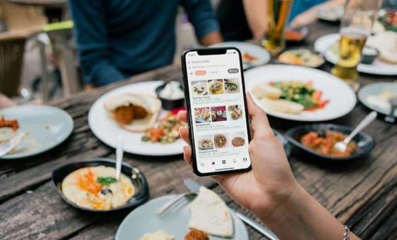 Hand holding smartphone displaying food delivery app at outdoor dining table with various dishes