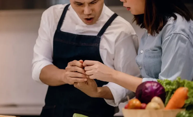 Chef in dark apron and woman in denim shirt examining injury in bright kitchen setting